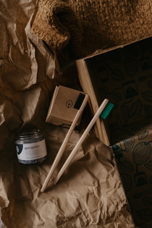 A rustic arrangement featuring eco-friendly toothbrushes made of wood and a jar of activated charcoal toothpaste. The items rest on crumpled brown paper, giving a natural and organic feel, with a background of cardboard and a textured fabric.