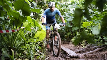 A cyclist wearing a blue uniform and a white helmet rides a mountain bike through a narrow, muddy trail surrounded by lush, green vegetation. The trail is uneven with visible roots, adding to the challenging terrain. The cyclist appears focused and determined, navigating through the thick jungle-like environment.
