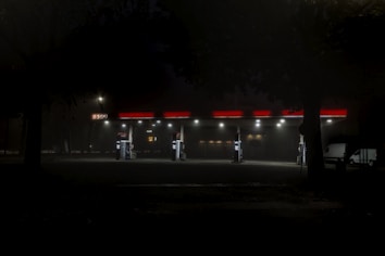 A dimly lit gas station at night with a foggy and mysterious atmosphere. The station has bright red accents on the roof and signs. Several gas pumps are visible, but the area is mostly empty with a couple of vehicles parked nearby. Trees frame the scene, adding to the eerie and quiet ambiance.