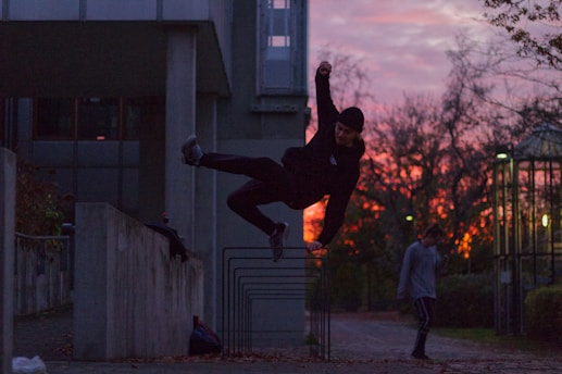 A person is performing a parkour move, leaping over a concrete wall in an urban setting as the sun sets, casting a warm glow in the sky. Another person walks in the background. The architecture and the trees contribute to the dramatic scene.