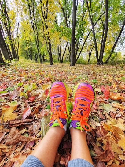 A pair of colorful running shoes rests on a carpet of autumn leaves in a forest. The trees are tall with a mix of green and yellow leaves, indicating the changing season. The perspective is from the point of view of someone sitting or lying down, looking up into the canopy.