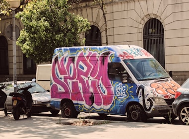 A van covered in colorful graffiti is parked on a city street. Vibrant pink, blue, and orange graffiti designs cover the vehicle, which is set against a backdrop of a stone building with large windows. Nearby, a scooter and several cars are also parked on the street alongside a tree.