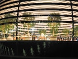 Model depicts the interior of a modern building, featuring several trees and seating areas. Multiple people are visible inside, engaged in various activities. The design of the building includes multiple horizontal lighted strips and a reflective glass facade that adds a dynamic feel.
