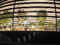 Model depicts the interior of a modern building, featuring several trees and seating areas. Multiple people are visible inside, engaged in various activities. The design of the building includes multiple horizontal lighted strips and a reflective glass facade that adds a dynamic feel.