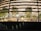 Model depicts the interior of a modern building, featuring several trees and seating areas. Multiple people are visible inside, engaged in various activities. The design of the building includes multiple horizontal lighted strips and a reflective glass facade that adds a dynamic feel.