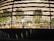 Model depicts the interior of a modern building, featuring several trees and seating areas. Multiple people are visible inside, engaged in various activities. The design of the building includes multiple horizontal lighted strips and a reflective glass facade that adds a dynamic feel.