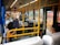 Inside a public bus, several passengers are seated, some wearing masks. The bus seats are blue with white head covers, and yellow poles and handles are visible. The bus appears to be in motion as the windows show blurred outdoor scenery.