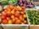 Various fruits and vegetables are displayed in wooden and wicker baskets. The foreground features a box of ripe, red tomatoes. Next to it is a box filled with small green citrus fruits. In the background, there are baskets containing green chayote and purple onions.