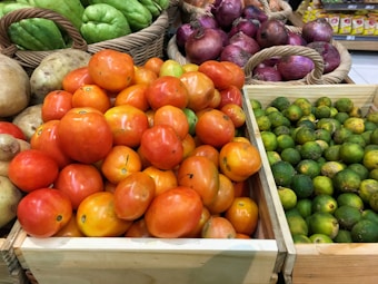Various fruits and vegetables are displayed in wooden and wicker baskets. The foreground features a box of ripe, red tomatoes. Next to it is a box filled with small green citrus fruits. In the background, there are baskets containing green chayote and purple onions.