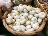 A large wicker basket filled with whole garlic bulbs, displayed in a market setting. The garlic bulbs are mostly white, with some discolorations, and have their stems intact. Other baskets and produce are visible in the background, including green peppers and potatoes.