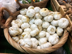 A large wicker basket filled with whole garlic bulbs, displayed in a market setting. The garlic bulbs are mostly white, with some discolorations, and have their stems intact. Other baskets and produce are visible in the background, including green peppers and potatoes.