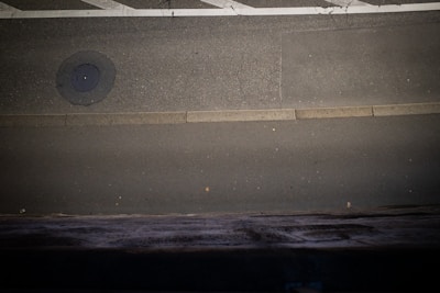 A paved road with visible cracks, featuring a manhole cover near the upper left and a light-colored curb running horizontally. The road is bordered by dark and textured surfaces at the bottom.