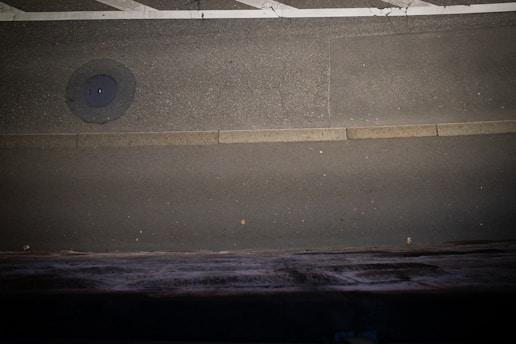 A paved road with visible cracks, featuring a manhole cover near the upper left and a light-colored curb running horizontally. The road is bordered by dark and textured surfaces at the bottom.