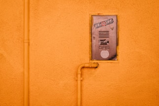 A gas meter cover is mounted on an orange stucco wall, featuring stickers and text. An orange pipe runs vertically alongside the cover and connects to it, adding an industrial element.