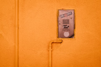 A gas meter cover is mounted on an orange stucco wall, featuring stickers and text. An orange pipe runs vertically alongside the cover and connects to it, adding an industrial element.