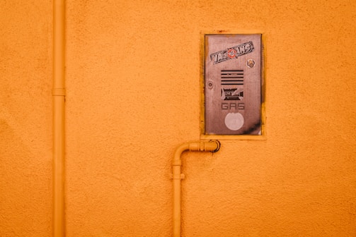 A gas meter cover is mounted on an orange stucco wall, featuring stickers and text. An orange pipe runs vertically alongside the cover and connects to it, adding an industrial element.