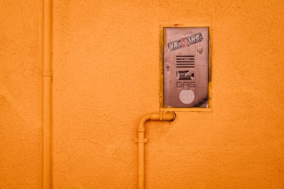 A gas meter cover is mounted on an orange stucco wall, featuring stickers and text. An orange pipe runs vertically alongside the cover and connects to it, adding an industrial element.