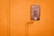 A gas meter cover is mounted on an orange stucco wall, featuring stickers and text. An orange pipe runs vertically alongside the cover and connects to it, adding an industrial element.