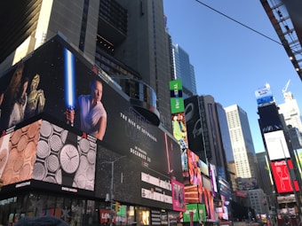 A bustling urban setting with numerous large digital billboards displaying various advertisements. One prominent advertisement features a movie poster for 'Star Wars: The Rise of Skywalker' with characters holding a lightsaber, while others include branding for watches and other products. The surrounding area is filled with tall skyscrapers reflecting daylight, indicating a commercial district.