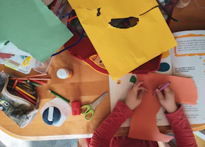 A child drawing on orange paper at a wooden table with craft supplies