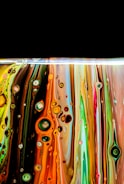 Colorful vertically flowing paint layers with circular patterns and bubbles, reminiscent of abstract art. The colors blend together in vibrant streaks, with a dramatic black background at the top creating contrast.