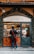 A person with a red sling bag stands in front of a traditional deli shop with a sign reading 'Antica Porchetteria Granieri 1916'. The shop window displays various bottles of wine and a selection of meats. Inside, a staff member wearing an apron and cap is attending to the customer.