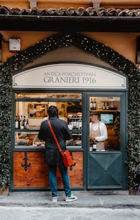 A person with a red sling bag stands in front of a traditional deli shop with a sign reading 'Antica Porchetteria Granieri 1916'. The shop window displays various bottles of wine and a selection of meats. Inside, a staff member wearing an apron and cap is attending to the customer.