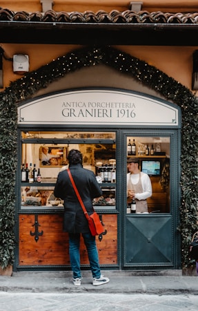 A person with a red sling bag stands in front of a traditional deli shop with a sign reading 'Antica Porchetteria Granieri 1916'. The shop window displays various bottles of wine and a selection of meats. Inside, a staff member wearing an apron and cap is attending to the customer.