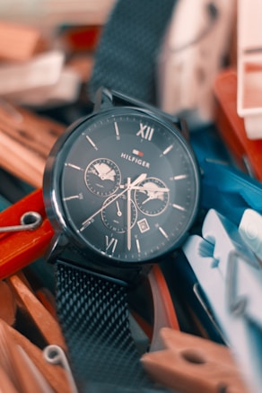 A wristwatch with a dark face and metallic mesh strap lays among a pile of colorful clothespins. The watch face displays Roman numerals and multiple sub-dials.