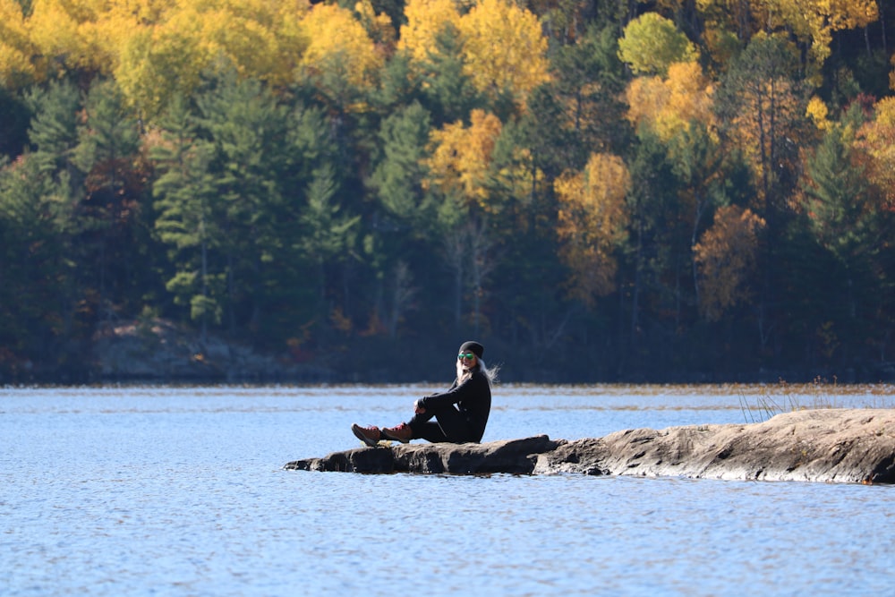 man in black jacket sitting on rock in the middle of the sea during daytime