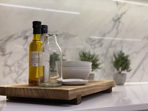 On a wooden cutting board, there's a bottle of olive oil, a glass bottle, stacked white bowls, and a small white dish. In the background, there are green potted plants on a white marble surface.
