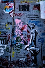 A concrete wall is covered with various graffiti and street art. There are colorful hearts, text messages like 'Positivity is the strongest currency' and 'Love', and a stenciled image of a child reaching upwards. The wall is layered with paint, featuring different styles and forms including doodles, symbols, and abstract shapes.