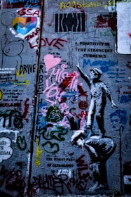 A concrete wall is covered with various graffiti and street art. There are colorful hearts, text messages like 'Positivity is the strongest currency' and 'Love', and a stenciled image of a child reaching upwards. The wall is layered with paint, featuring different styles and forms including doodles, symbols, and abstract shapes.