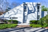 A modern, minimalist white house with a unique angular roof design. The facade features large windows and a simple door. Shadows of tree branches are cast on the front, adding an artistic touch. Lush green bushes and shrubs surround the exterior, providing a contrast to the clean lines of the architecture. The background shows a clear blue sky and bare trees.