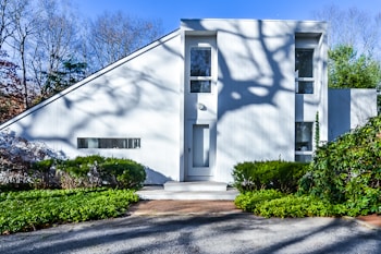 A modern, minimalist white house with a unique angular roof design. The facade features large windows and a simple door. Shadows of tree branches are cast on the front, adding an artistic touch. Lush green bushes and shrubs surround the exterior, providing a contrast to the clean lines of the architecture. The background shows a clear blue sky and bare trees.