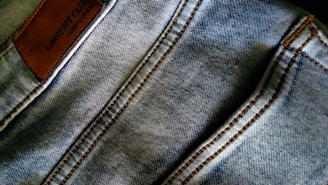 Denim fabric with visible stitching lines in a light blue wash. A brown leather patch with embossed text is located on the upper left area.