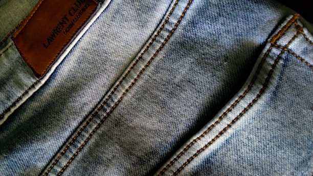 Denim fabric with visible stitching lines in a light blue wash. A brown leather patch with embossed text is located on the upper left area.