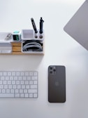 A neatly organized workspace featuring a wireless keyboard, a black smartphone, a wooden desk organizer with compartments for pens and other items, and part of a laptop. The desk is clean and minimalistic.