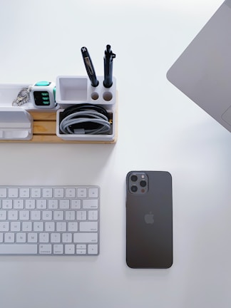 A neatly organized workspace featuring a wireless keyboard, a black smartphone, a wooden desk organizer with compartments for pens and other items, and part of a laptop. The desk is clean and minimalistic.