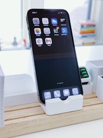 A smartphone is placed upright in a white docking station on a wooden surface. The screen displays several app icons and the time at the top left corner. In the background, there are blurred colorful books stacked and additional white containers.