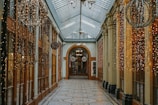 An ornate indoor passageway is decorated with cascading strings of small, warm white fairy lights on both sides. The ceiling is made of glass, allowing diffuse natural light to fill the space. The walls are lined with large wooden-framed windows and doors, while the floor features a patterned surface. Decorative spherical light fixtures hang from above, enhancing the festive atmosphere.