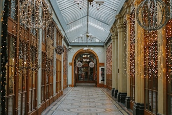 An ornate indoor passageway is decorated with cascading strings of small, warm white fairy lights on both sides. The ceiling is made of glass, allowing diffuse natural light to fill the space. The walls are lined with large wooden-framed windows and doors, while the floor features a patterned surface. Decorative spherical light fixtures hang from above, enhancing the festive atmosphere.