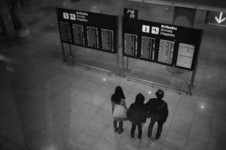 Three people stand together in front of a large airport arrivals board, checking flight information displayed on digital screens. Overhead, the lighting casts reflections on the tiled floor, and there is a visible arrow sign indicating direction.