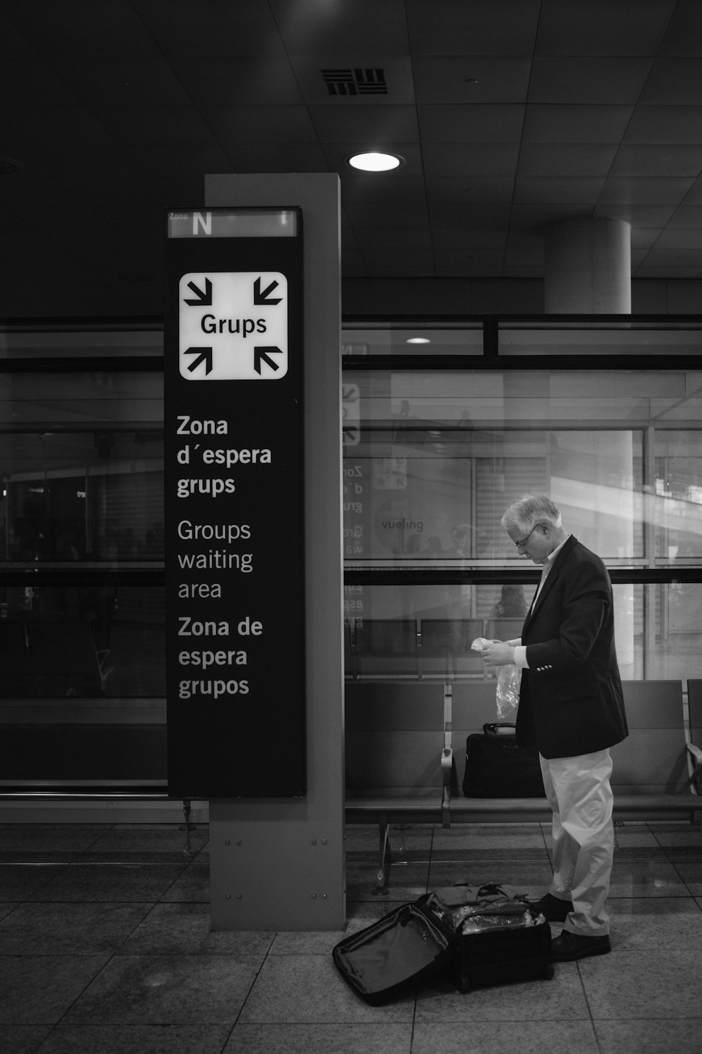 Photo en niveaux de gris d’un homme en veste noire et pantalon blanc debout à côté d’une fenêtre en verre