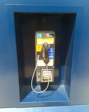 A traditional payphone is mounted on a blue wall. It features a handset attached by a coiled cord, a coin slot, and various instructional stickers in different colors including yellow and blue.