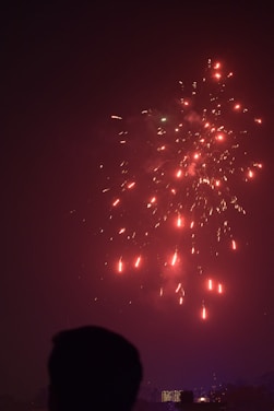 Fireworks light up the night sky with bright red and yellow sparks. A silhouetted figure in the foreground suggests a human presence, watching the display. Below, the skyline features illuminated buildings, hinting at an urban setting.