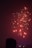 Fireworks light up the night sky with bright red and yellow sparks. A silhouetted figure in the foreground suggests a human presence, watching the display. Below, the skyline features illuminated buildings, hinting at an urban setting.