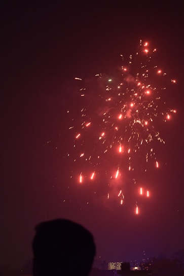 Fireworks light up the night sky with bright red and yellow sparks. A silhouetted figure in the foreground suggests a human presence, watching the display. Below, the skyline features illuminated buildings, hinting at an urban setting.