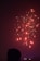 Fireworks light up the night sky with bright red and yellow sparks. A silhouetted figure in the foreground suggests a human presence, watching the display. Below, the skyline features illuminated buildings, hinting at an urban setting.