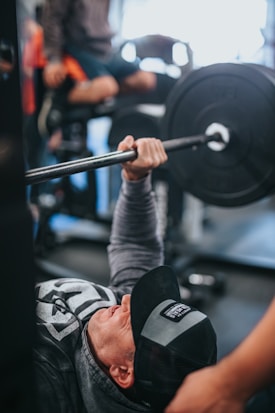 A person wearing a cap and sweatshirt is performing a bench press in a gym setting. The focus is on the effort and concentration of the individual lifting a heavy barbell, with another person nearby offering support or guidance. The gym environment is slightly blurred in the background, highlighting fitness equipment.